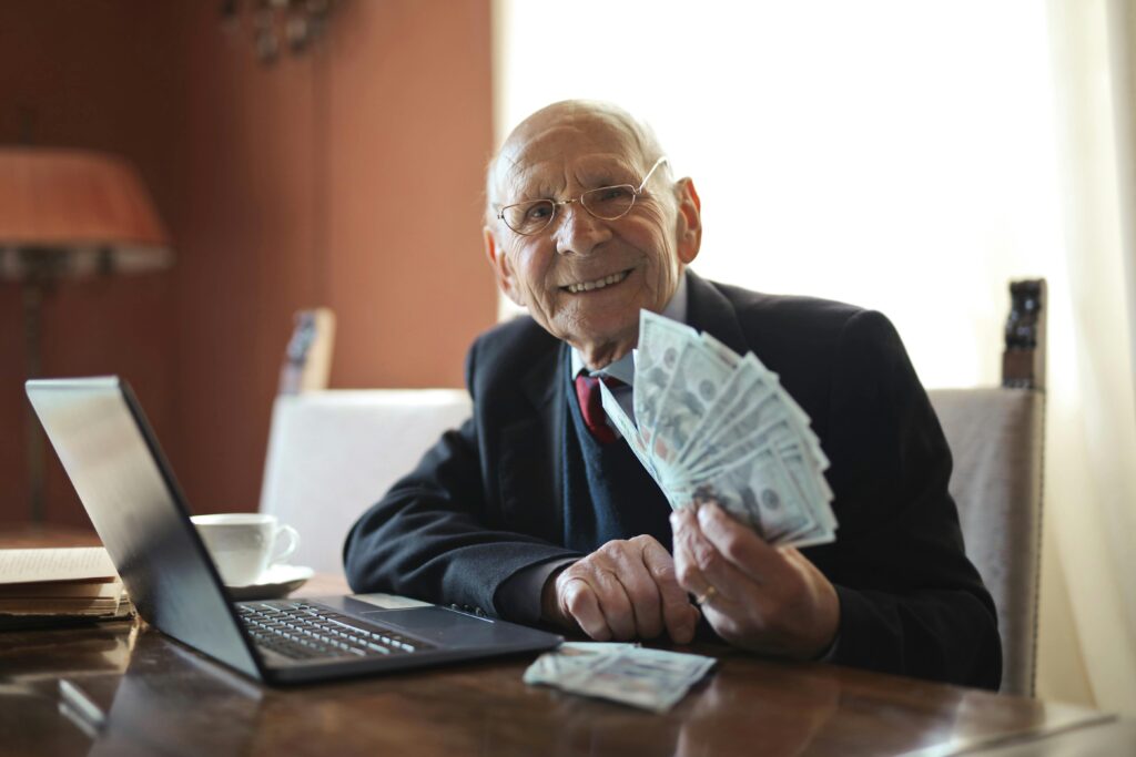 Earn 30% With Revglobal Happy senior businessman showing cash at his desk with a laptop, indoors.